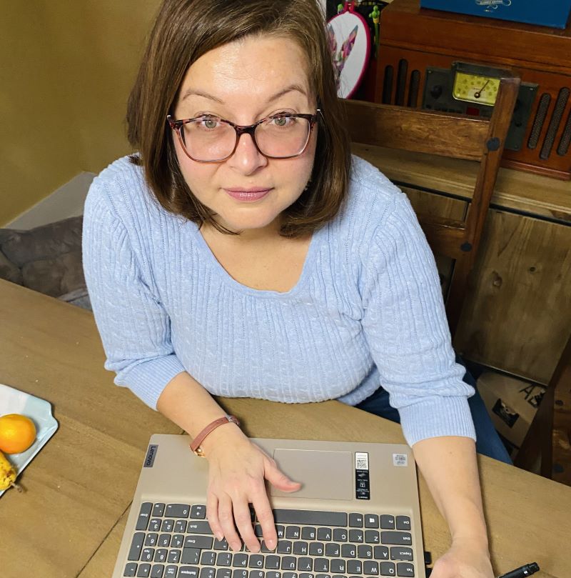 Photo taken from above Dinah, who is seated in front of her laptop. She is looking up into the camera. She has brown hair, shoulder-length, with glasses.